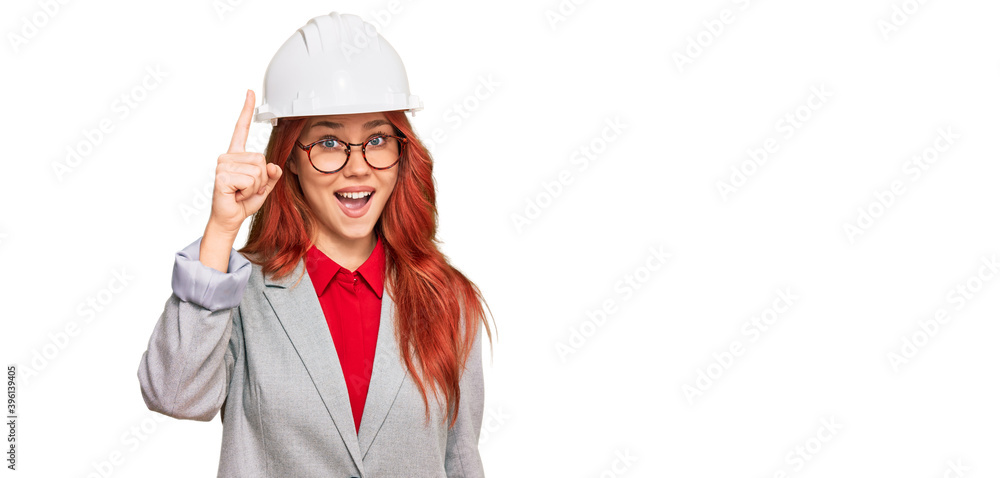 Young redhead woman wearing architect hardhat pointing finger up with successful idea. exited and happy. number one. Young redhead woman wearing architect hardhat pointing finger up with successful idea. exited and happy. number one.