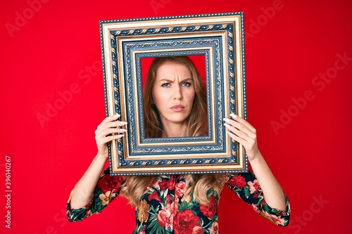 Young caucasian woman with blond hair holding empty frame skeptic and nervous, frowning upset because of problem. negative person.
