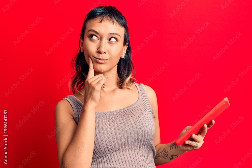 Naklejka premium Young woman holding touchpad tablet serious face thinking about question with hand on chin, thoughtful about confusing idea