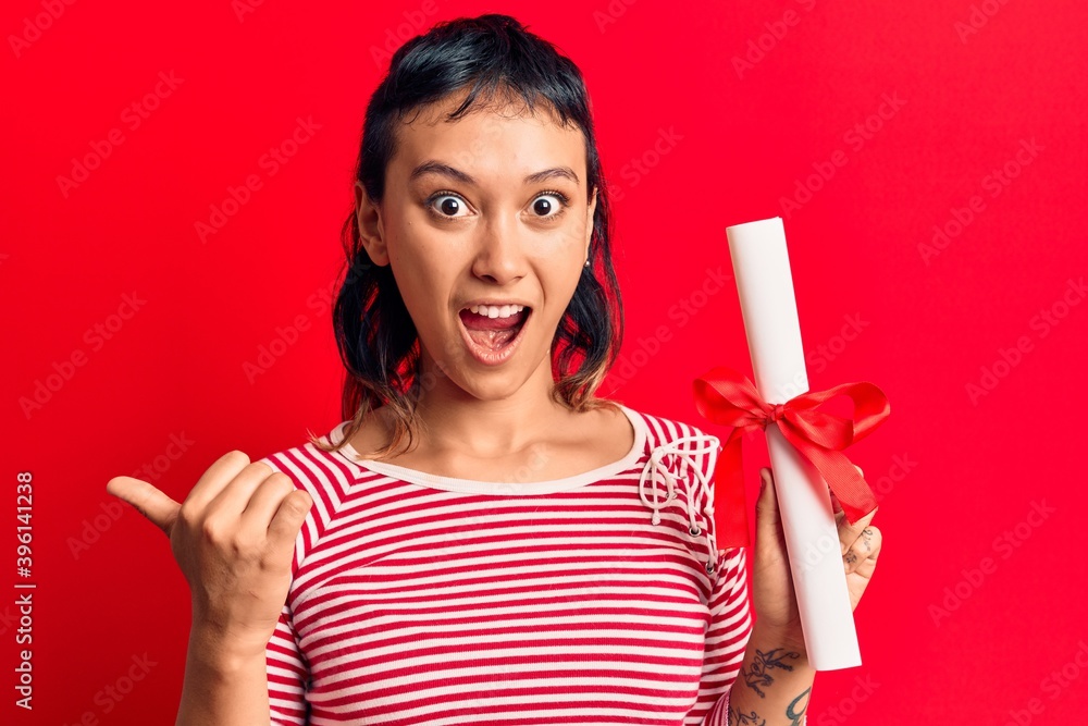 Young woman holding graduate degree diploma pointing thumb up to the side smiling happy with open mouth Young woman holding graduate degree diploma pointing thumb up to the side smiling happy with open mouth