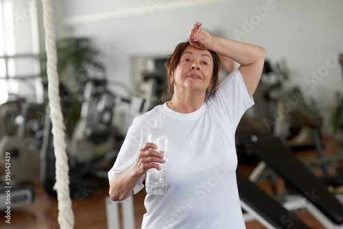 Tired senior woman after gym workout. Exhausted woman wiping sweat while holding bottle of water at fitness center.