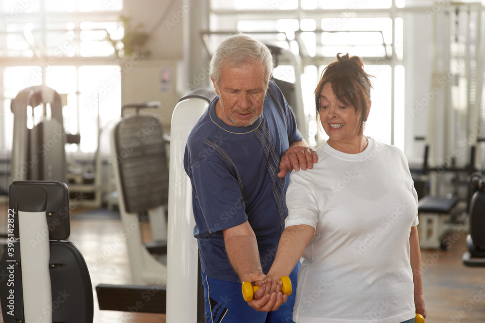 Fototapeta premium Senior woman lifting dumbbell at gym. Older trainer helping to woman lifting dumbbell at fitness club. People, retirement, leisure.