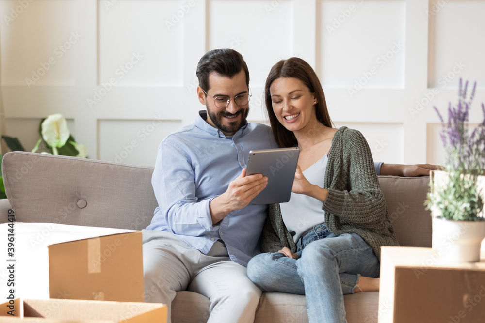 Smiling young Caucasian couple relax on sofa in living room on moving day watch video on tablet. Happy man and woman renters tenants rest on couch relocate to new home, use pad browsing internet. Smiling young Caucasian couple relax on sofa in living room on moving day watch video on tablet. Happy man and woman renters tenants rest on couch relocate to new home, use pad browsing internet.