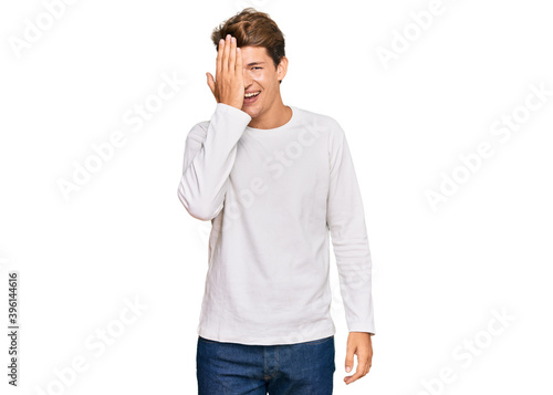 Handsome caucasian man wearing casual white sweater covering one eye with hand, confident smile on face and surprise emotion.