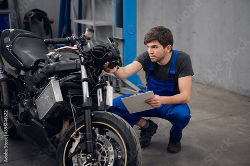 A workman inspects a motorcycle. He is wearing a blue overalls