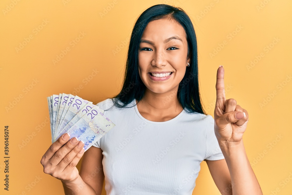 Beautiful hispanic woman holding 20 swedish krona banknotes smiling with an idea or question pointing finger with happy face, number one Beautiful hispanic woman holding 20 swedish krona banknotes smiling with an idea or question pointing finger with happy face, number one