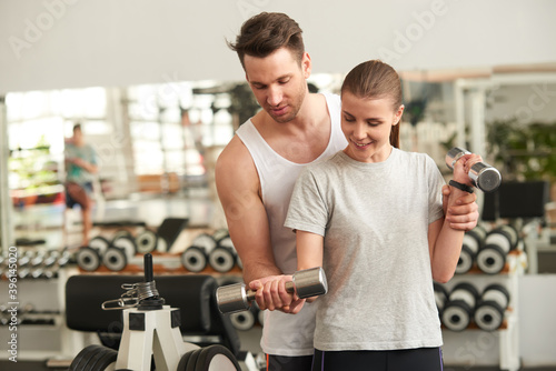 Young woman lifting dumbbells with personal trainer. Young handsome trainer helping woman lifting weights in fitness room. Young couple exercise together in gym.