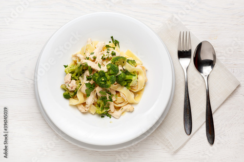 Pasta with broccoli and chicken on a white plate on a white wooden table with a spoon and fork
