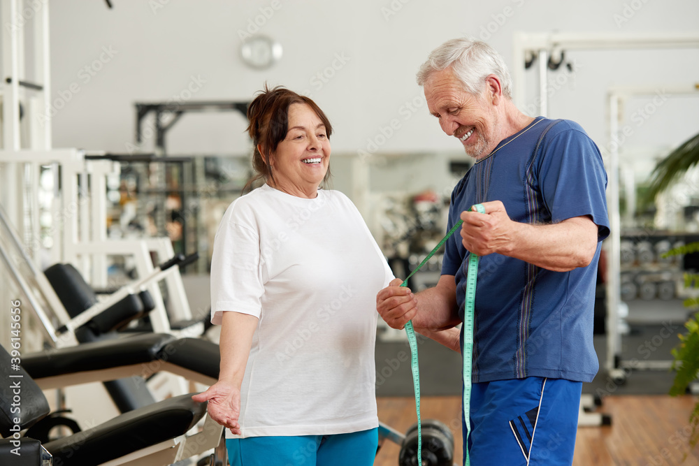 Senior woman excited about her weight loss. Happy mature couple with measuring tape at gym. Woman satisfied with her weight loss progress. Great fitness results concept. Senior woman excited about her weight loss. Happy mature couple with measuring tape at gym. Woman satisfied with her weight loss progress. Great fitness results concept.