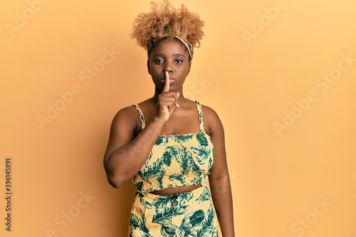 Young african woman with afro hair wearing summer dress asking to be quiet with finger on lips. silence and secret concept.