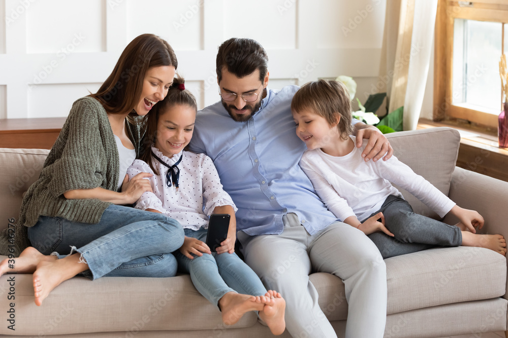 Overjoyed young Caucasian parents with two small kids relax on comfortable couch at home using smartphone. Happy family with children relax on sofa in living room watching video on cellphone gadget.