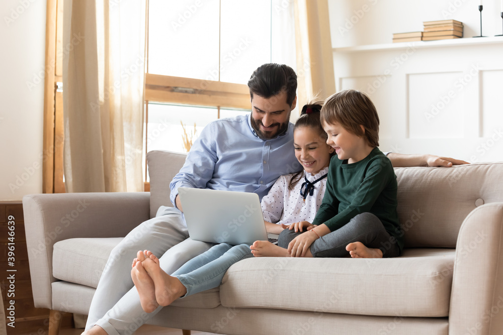 Caring Caucasian dad and little children sit relax on sofa at home using modern laptop together. Happy loving young father enjoy weekend with two kids watch video browse internet on computer.