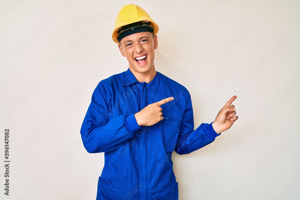 Young hispanic boy wearing worker uniform and hardhat smiling and looking at the camera pointing with two hands and fingers to the side.