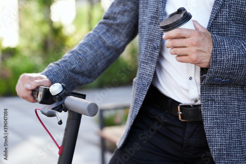 Necessary things. Close up of hands of stylish middle aged businessman holding a cup of coffee while standing with his electrical scooter outdoors in the daytime