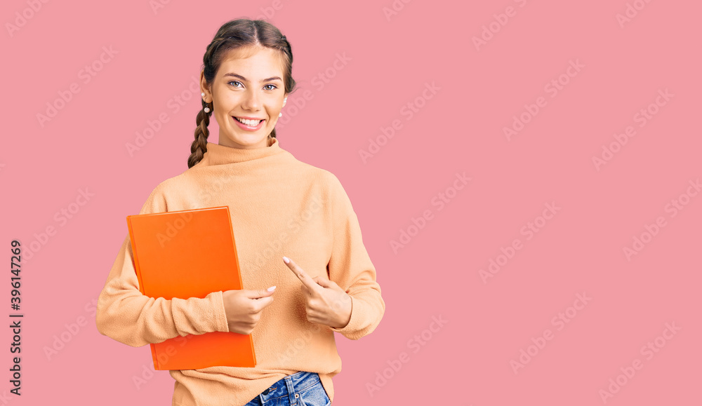 Beautiful caucasian woman with blonde hair holding book smiling happy pointing with hand and finger Beautiful caucasian woman with blonde hair holding book smiling happy pointing with hand and finger
