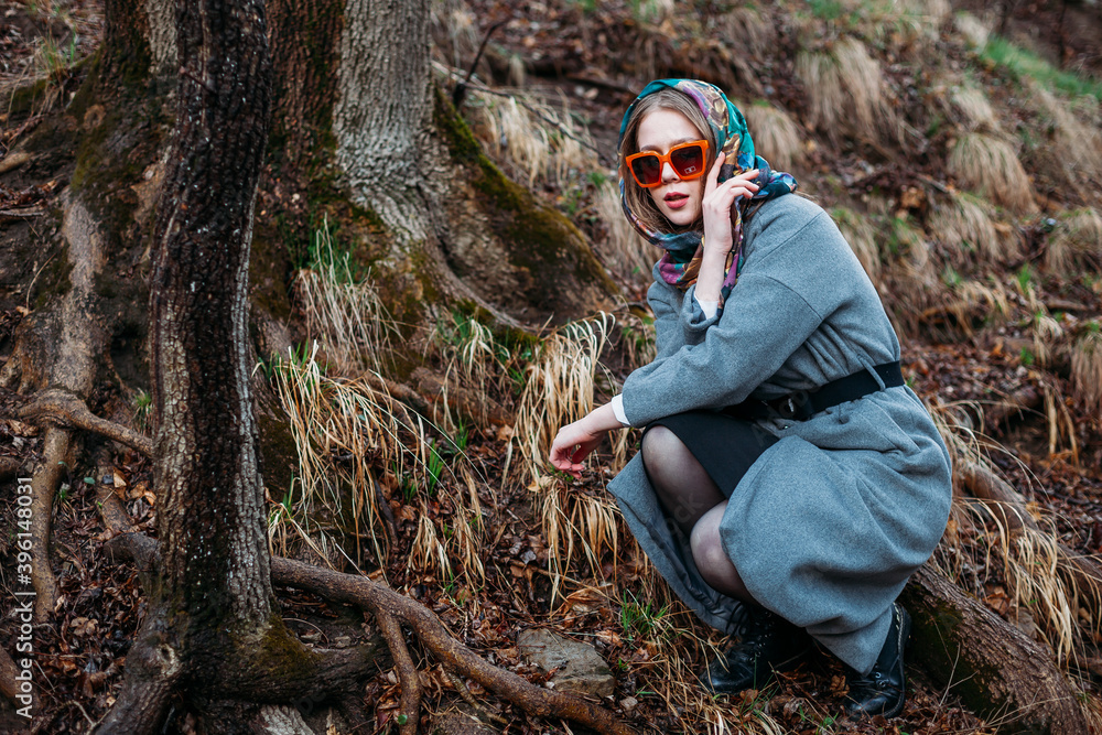 Naklejka premium fashion shooting. young model wearing bright silk headscarf and orange-rimmed sunglasses sits under tree in an autumn gray forest