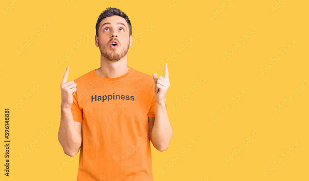 Handsome young man with bear wearing tshirt with happiness word message amazed and surprised looking up and pointing with fingers and raised arms. Handsome young man with bear wearing tshirt with happiness word message amazed and surprised looking up and pointing with fingers and raised arms.