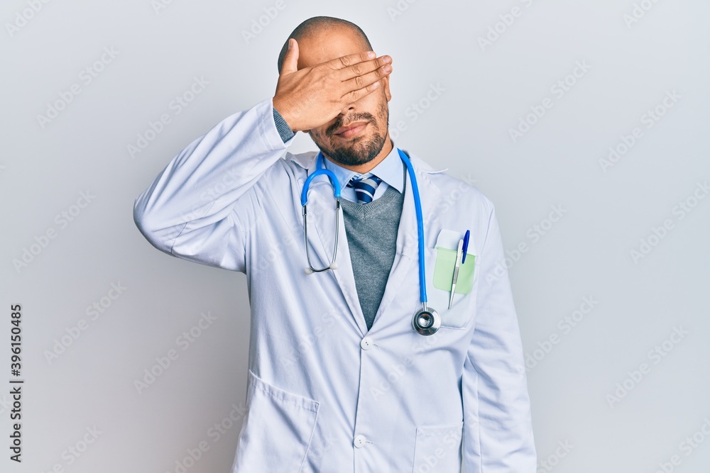 Hispanic adult man wearing doctor uniform and stethoscope covering eyes with hand, looking serious and sad. sightless, hiding and rejection concept Hispanic adult man wearing doctor uniform and stethoscope covering eyes with hand, looking serious and sad. sightless, hiding and rejection concept