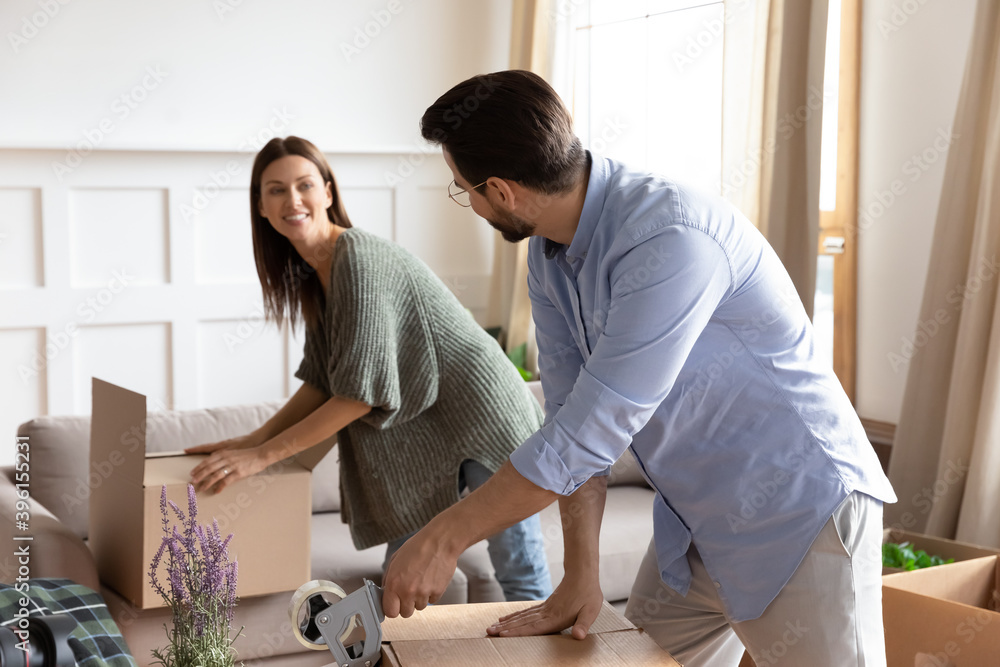 Fototapeta premium Happy young couple renters packing on moving day wrap packages parcels with adhesive scotch in living room. Excited Caucasian family tenants relocate seal boxes with tape. Delivery concept.