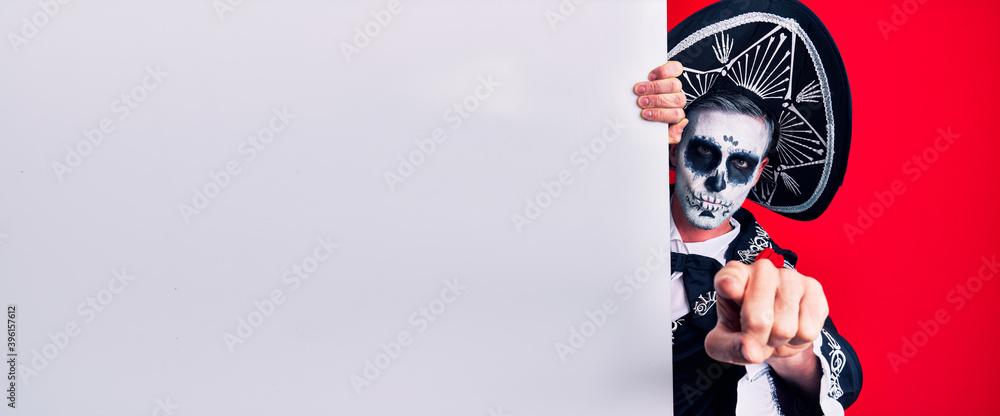Young man wearing mexican day of the dead costume holding blank empty banner pointing with finger to the camera and to you, confident gesture looking serious Young man wearing mexican day of the dead costume holding blank empty banner pointing with finger to the camera and to you, confident gesture looking serious
