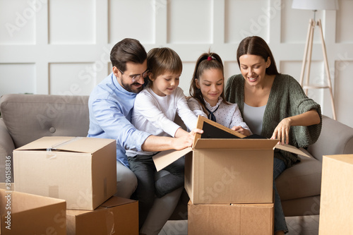 Excited young Caucasian family with two kids have fun unboxing packages relocate to new home together. Happy parents with small children unpack boxes moving to own house. Real estate concept.