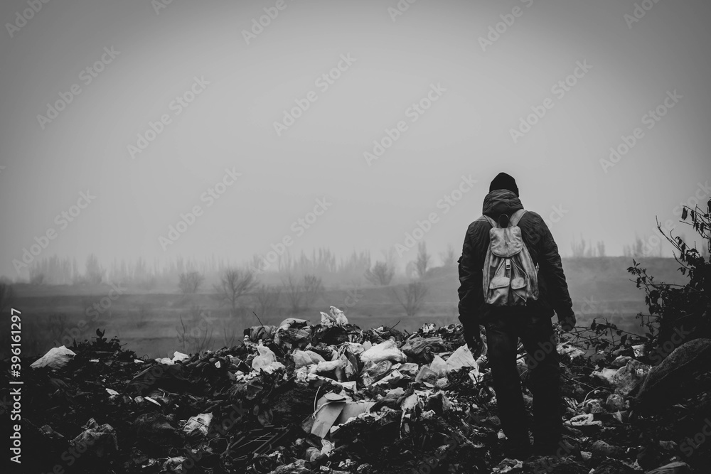 A man in old clothes and with a backpack stands against the background of a landfill.