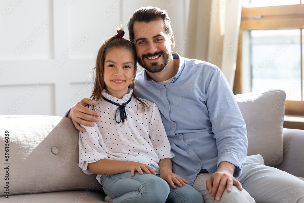 Portrait of happy young Caucasian dad and little schoolgirl daughter sit rest on sofa in living room hugging. Smiling single father embrace small girl child show love care, relax together in new home. Portrait of happy young Caucasian dad and little schoolgirl daughter sit rest on sofa in living room hugging. Smiling single father embrace small girl child show love care, relax together in new home.