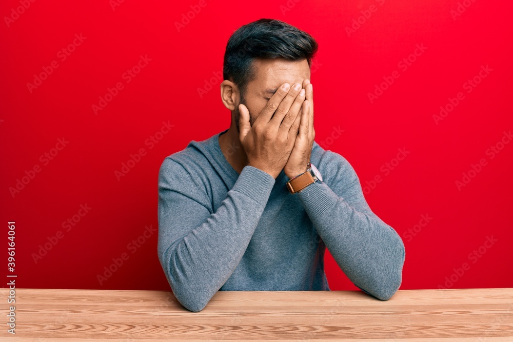 Handsome hispanic man wearing casual style sitting on the table with sad expression covering face with hands while crying. depression concept. Handsome hispanic man wearing casual style sitting on the table with sad expression covering face with hands while crying. depression concept.