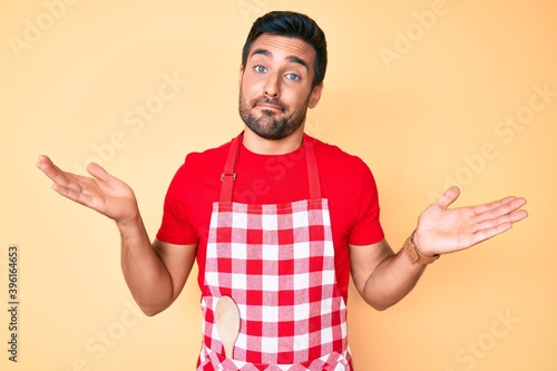 Young hispanic man wearing professional baker apron clueless and confused with open arms, no idea and doubtful face.