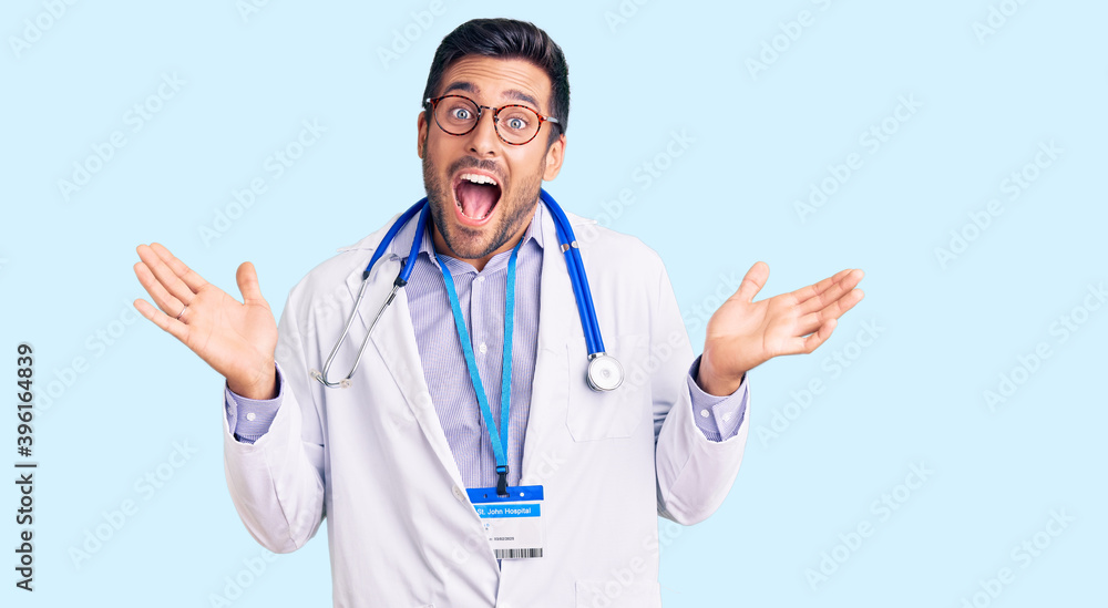 Young hispanic man wearing doctor uniform and stethoscope celebrating crazy and amazed for success with arms raised and open eyes screaming excited. winner concept Young hispanic man wearing doctor uniform and stethoscope celebrating crazy and amazed for success with arms raised and open eyes screaming excited. winner concept