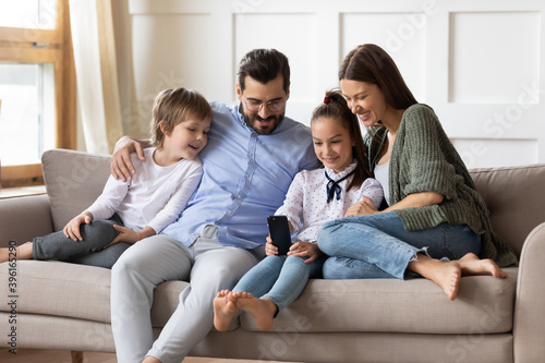 Happy young Caucasian family with two small kids sit relax on sofa in living room watch funny video on cellphone. Smiling parents with children rest on couch at home using smartphone on weekend.