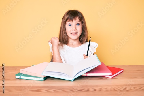 Little caucasian kid girl with long hair studying for school exam pointing thumb up to the side smiling happy with open mouth