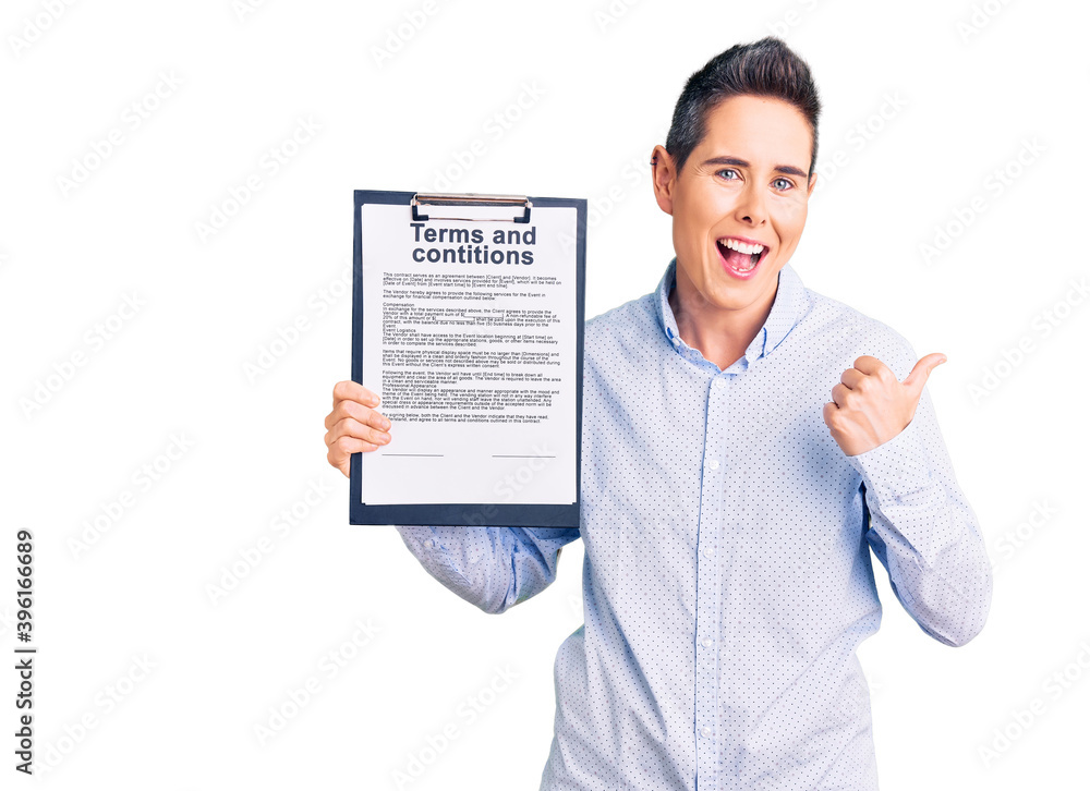 Young woman with short hair holding clipboard with terms and conditions document pointing thumb up to the side smiling happy with open mouth Young woman with short hair holding clipboard with terms and conditions document pointing thumb up to the side smiling happy with open mouth