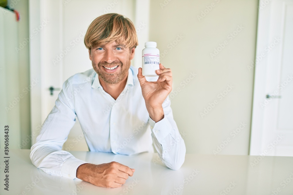 Young irish man smiling happy holding bottle with pills sitting on the table at home. Young irish man smiling happy holding bottle with pills sitting on the table at home.