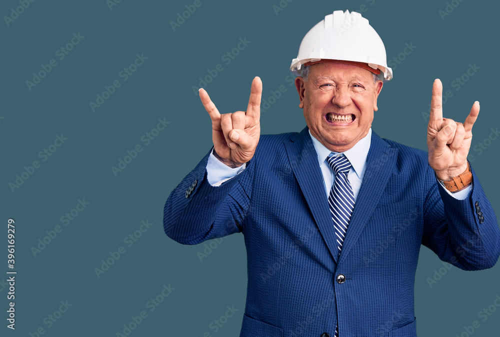 Senior handsome grey-haired man wearing suit and architect hardhat shouting with crazy expression doing rock symbol with hands up. music star. heavy concept. Senior handsome grey-haired man wearing suit and architect hardhat shouting with crazy expression doing rock symbol with hands up. music star. heavy concept.