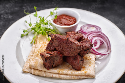shashlik, with sauce, onions, and lavash, on a white plate, on a dark background