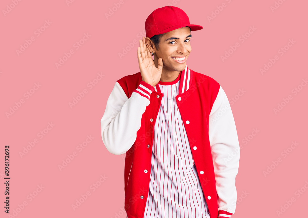 Young african amercian man wearing baseball uniform smiling with hand over ear listening an hearing to rumor or gossip. deafness concept. Young african amercian man wearing baseball uniform smiling with hand over ear listening an hearing to rumor or gossip. deafness concept.