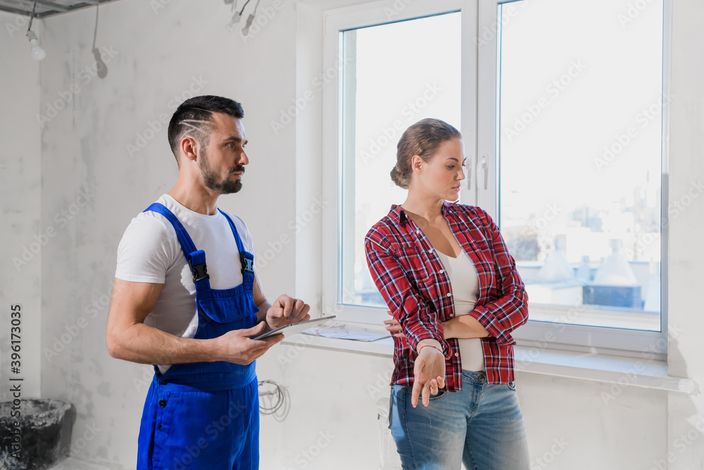 A builder in a blue overalls shows the client the renovation of an apartment. He writes notes with a pen