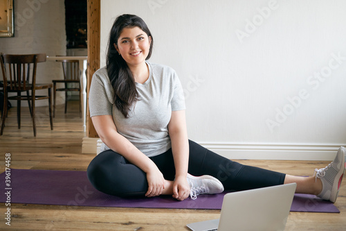 Indoor image of beautiful energetic young plus size woman with curvy body training at home to lose weight and get strong flexible body, wearing sneakers, exercising on mat, watching video on laptop photo