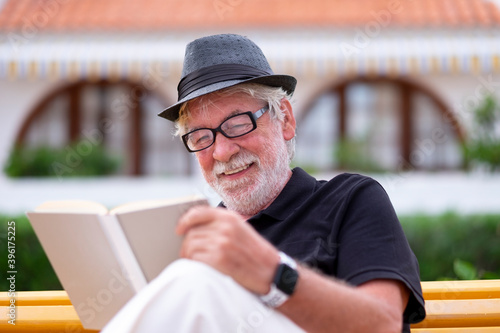 Portrait of a senior bearded man with hat sitting on a bench in the park relaxing and reading a book. Smiling carefree people during retirement