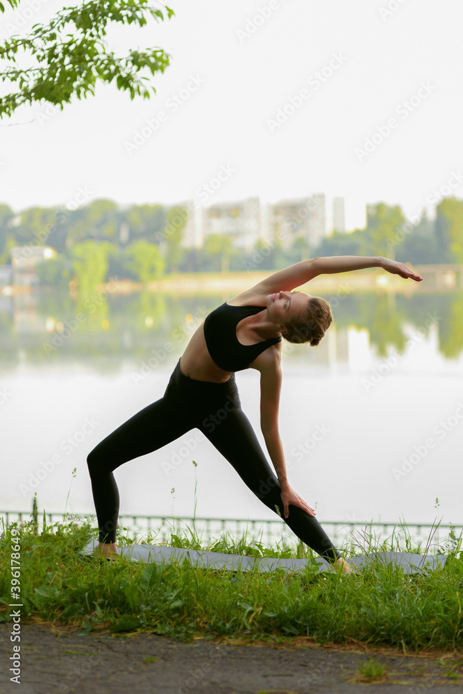 Fototapeta premium Full length image of a young woman in black sportwear doing yoga in the park, lake background.