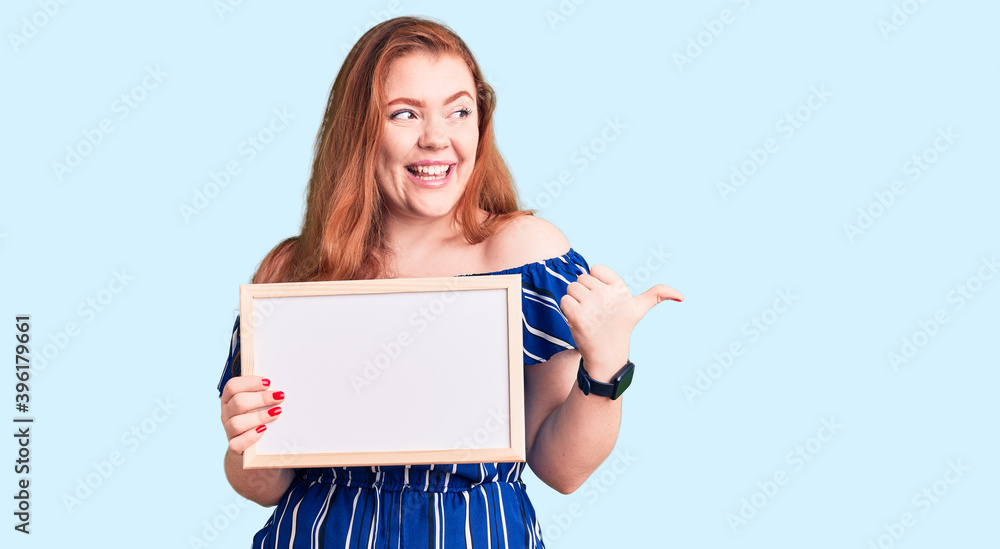 Naklejka premium Young beautiful redhead woman holding empty white chalkboard pointing thumb up to the side smiling happy with open mouth