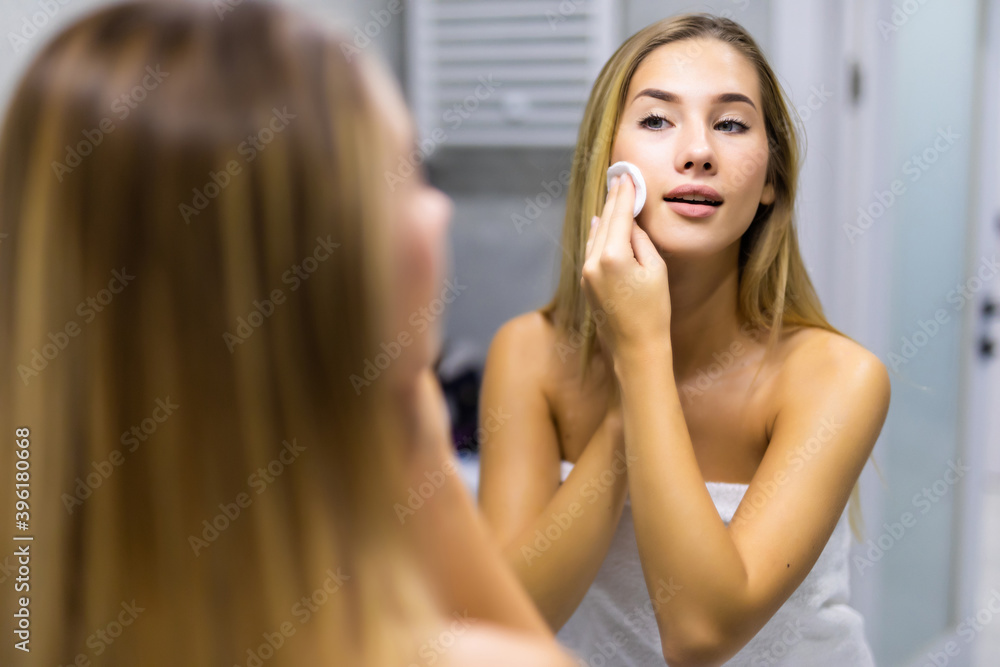 Fototapeta premium Beauty, skin care and people concept - smiling young woman applying lotion to cotton disc for washing her face at bathroom