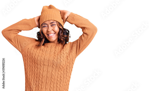 Young beautiful mixed race woman wearing wool sweater and winter hat relaxing and stretching, arms and hands behind head and neck smiling happy photo