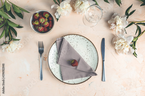 Flat lay composition with white peony flowers, empty plate with napkin and cutlery on a light pink concrete background