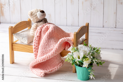 Teddy bear is lying in a small wooden bed, covered with a knitted blanket. Next to it is a pot with an artificial flower. Baby sleep concept photo