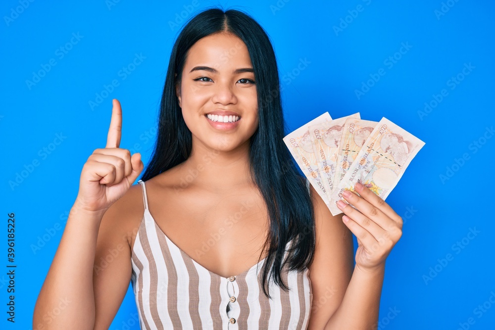 Young beautiful asian girl holding united kingdom pounds smiling with an idea or question pointing finger with happy face, number one Young beautiful asian girl holding united kingdom pounds smiling with an idea or question pointing finger with happy face, number one