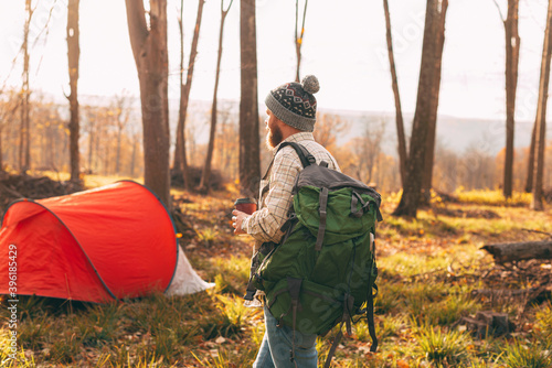 Bearded man with cup of coffee, wearing backgack near red tent at forest outdoor.