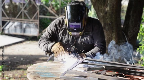 Man welding some irons on a wooden table, protected with a mask and gloves, in an outdoor space with trees