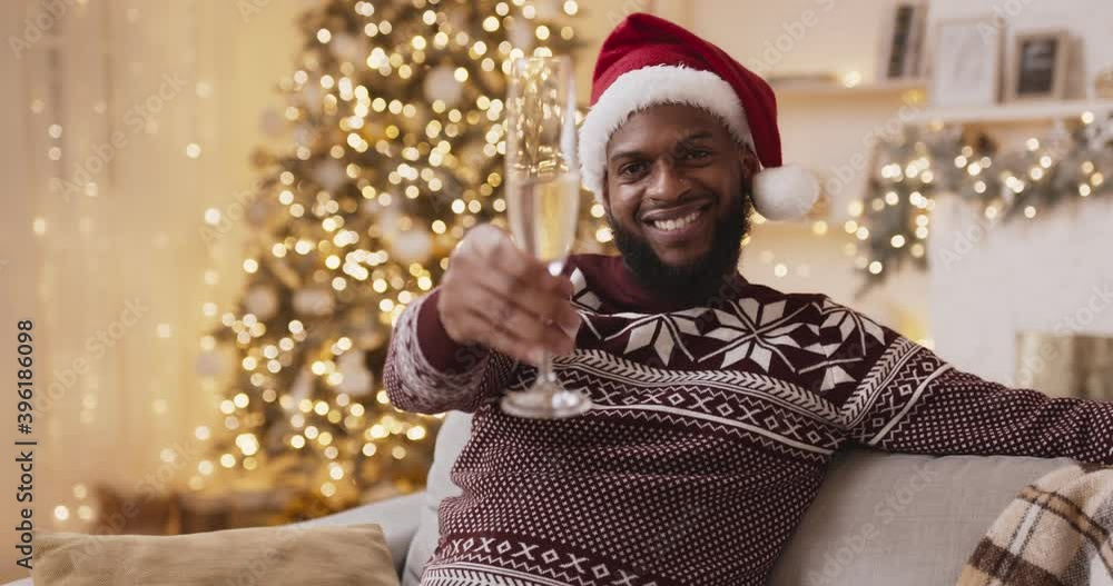 Young african american man in winter sweater and santa hat clinking to camera with champagne Young african american man in winter sweater and santa hat clinking to camera with champagne